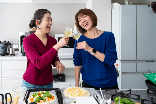 Happy Asian women clinking wine glasses together while making homemade pizzas in a bright kitchen