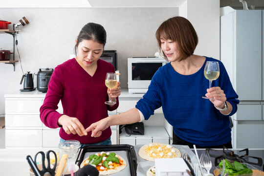 Two Asian women holding wine glasses and adding fresh toppings to homemade pizzas at a kitchen counter