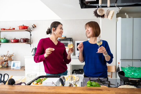 Two happy Asian women holding wine glasses and dancing playfully while making homemade pizzas in a bright kitchen