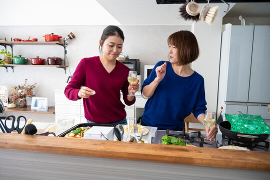 Two happy Asian women holding wine glasses and dancing playfully while making homemade pizzas in a bright kitchen