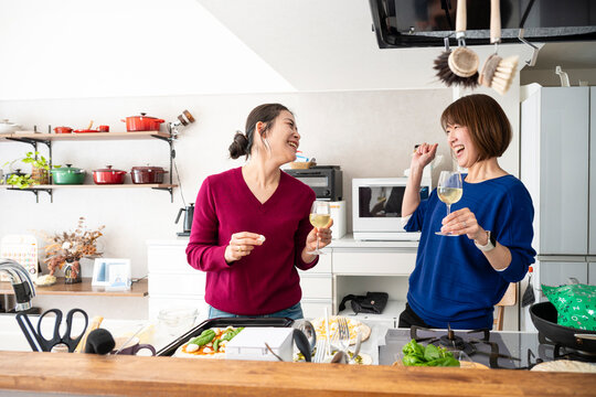 Two cheerful Asian women laughing heartily and holding wine glasses in a modern kitchen