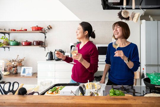 Two smiling Asian women holding wine glasses and looking away together in a bright kitchen