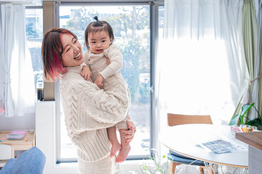 Happy Asian mother holding up her sweet toddler and smiling together in a sunlit room