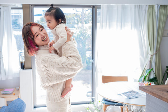Happy Asian mother holding up her sweet toddler and smiling together in a sunlit room