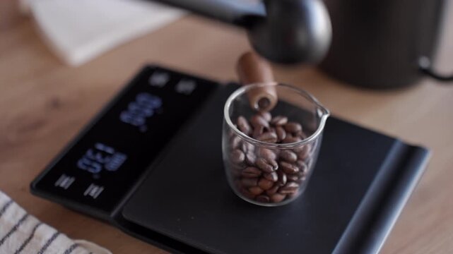 Coffee beans pouring with scoop in glass cup for measuring on digital scale before brewing. Concept of coffee preparation and home brewing
