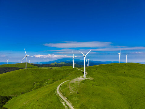 Wind turbines stand on rolling green hills under a bright blue sky with some clouds in the distance on a sunny day