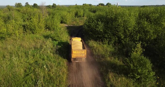 Aerial tracking shot following a yellow dump truck driving on a dirt road through lush green countryside