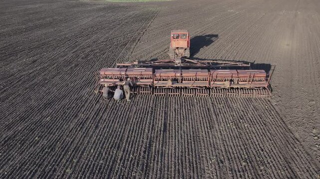 Farm workers repairing old seed drill in cultivated field during sowing season, agricultural machinery maintenance and equipment repair from aerial drone view