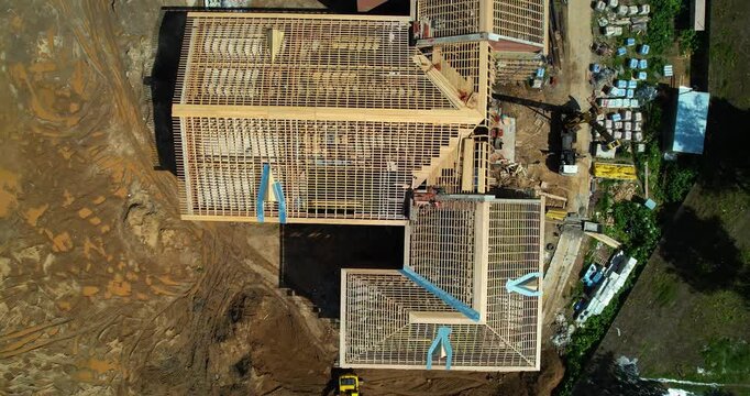 Aerial top-down tracking shot of a building under construction showing wooden roof trusses and a yellow excavator working nearby