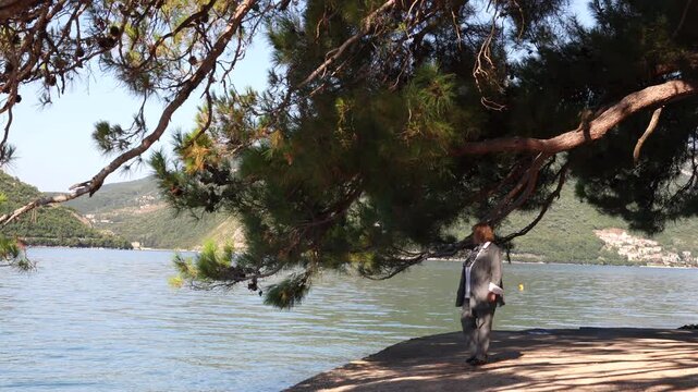 Senior Woman Walking by the Sea Under Pine Trees. Senior woman walking near the sea under pine trees with coastal mountains in the background. Peaceful Mediterranean travel and outdoor lifestyle scene