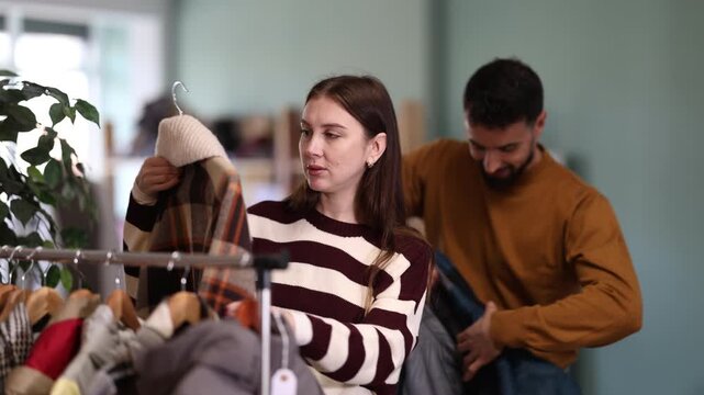 Elegance european couple shopping clothes, young woman choosing coat or down jacket and young man trying new down jacket for cold season in modern store