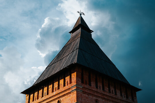 A tall brick tower with a pointed roof stands prominently against a dramatic sky filled with clouds