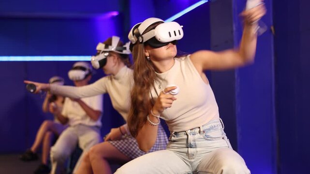 Group of excited young girls sitting with handheld controllers in hands engaged in virtual world experiencing through VR glasses covering face. Concept of immersion and interactivity in game