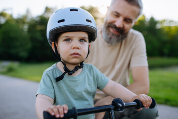 Naklejka premium Father teaching toddler son to ride balance bike while wearing safety helmet.