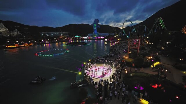 Night aerial view of amusement park lake light show with crowd and roller coaster