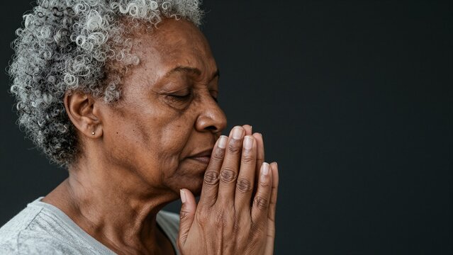 Close-up of an elderly African woman with curly gray hair, eyes closed in prayer, hands clasped in devotion. She appears deeply serene and connected spiritually against a dark background.