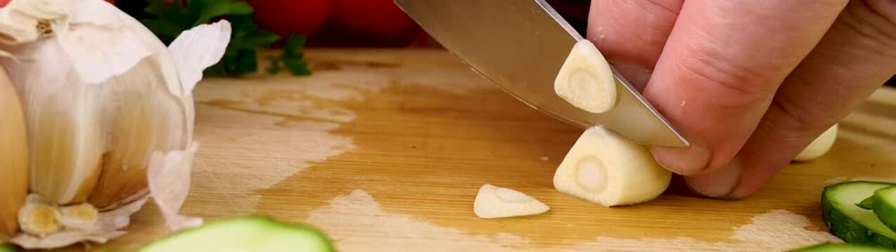 Hand using a sharp metal knife to meticulously slice a peeled garlic clove on a wooden board during food preparation.