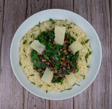 A bowl contains a serving of mushroom and spinach risotto. On top are pieces of Morteau sausage and parmesan cheese. The dish is placed on a wooden surface.