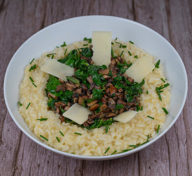 A bowl contains a serving of mushroom and spinach risotto. On top are pieces of Morteau sausage and parmesan cheese. The dish is placed on a wooden surface.