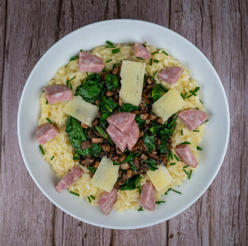 A bowl contains a serving of mushroom and spinach risotto. On top are pieces of Morteau sausage and parmesan cheese. The dish is placed on a wooden surface.