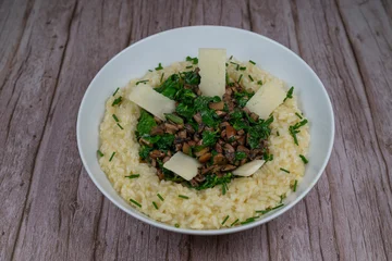 A bowl contains a serving of mushroom and spinach risotto. On top are pieces of Morteau sausage and parmesan cheese. The dish is placed on a wooden surface. © FreeProd