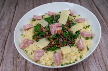 A bowl contains a serving of mushroom and spinach risotto. On top are pieces of Morteau sausage and parmesan cheese. The dish is placed on a wooden surface. © FreeProd