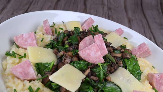A bowl contains a serving of mushroom and spinach risotto. On top are pieces of Morteau sausage and parmesan cheese. The dish is placed on a wooden surface.
