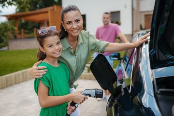 Obraz premium Mother and girl charging electric car while family prepares for summer trip.
