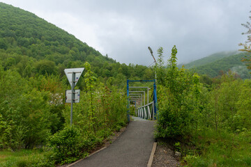 Fototapeta premium Steel footbridge and walking path cross a green valley surrounded by forested mountains. Foggy hills and dense vegetation create a scenic hiking environment.