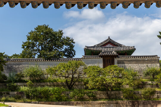 Traditional Korean palace pavilion and historic stone wall on a green terraced hill under a cloudy blue sky.