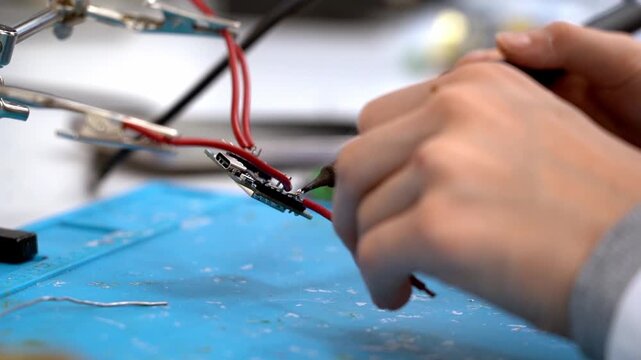 Hands hold wires and soldering iron. Focus on precise, careful work on circuitry. Blue mat shows workspace clutter. Detail reveals technical skill and concentration. Ideal for electronics, DIY