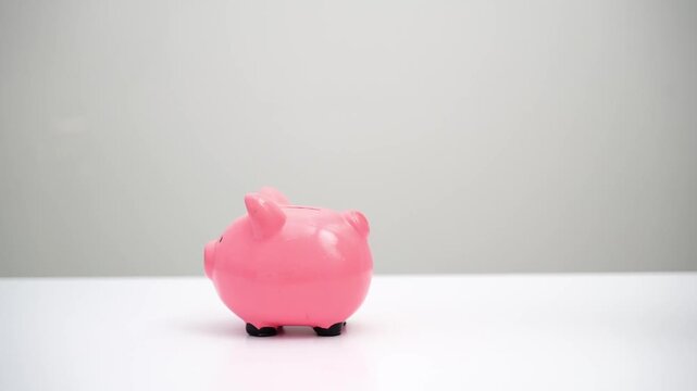Pink piggy bank shatters on white surface. Coins spill everywhere as hammer strikes. Hand holds tool with deliberate force. Scene conveys financial loss or breaking of savings