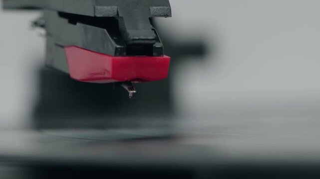Red needle rests on spinning vinyl record. Black stylus hovers just above groove. Music fades as needle gently begins to trace. Close-up captures vinyl playback moment