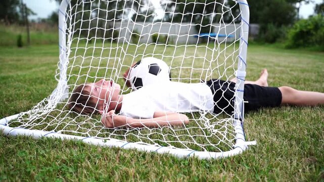 Young boy rests inside soccer goal, smiling. He lies on grass, head tilted back, eyes closed. Black and white ball rests near his cheek. Bright green grass surrounds the white net