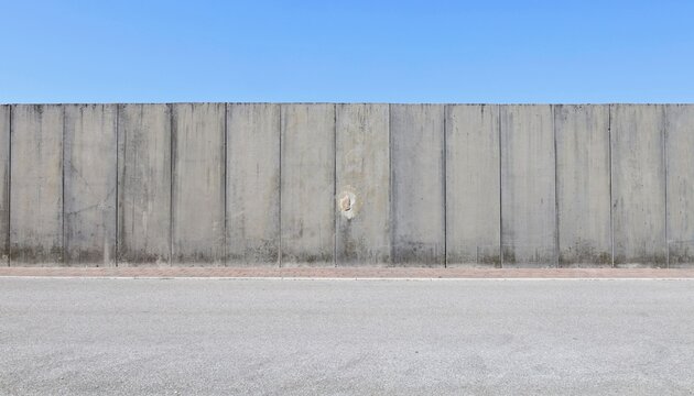 High prefab concrete boundary wall with blue sky above. Red tled sidewalk and street in front. Background for copy space.