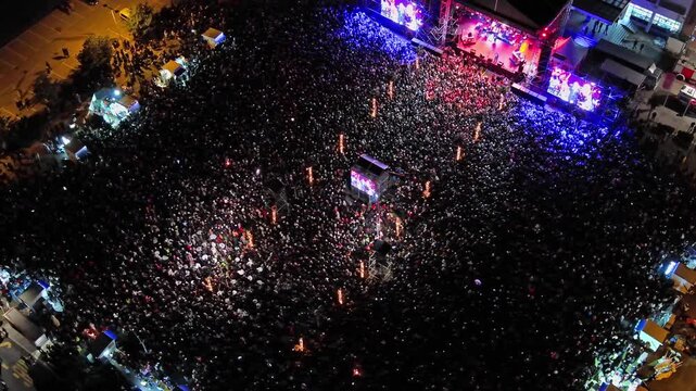 Aerial around view of crowd at night music concert. People gathered at festival outdoor stage with lights and screens, 4k