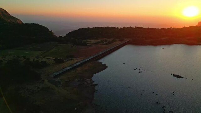 aerial drone view of a vibrant golden sunset over the reservoir of Ghatghar Dam and the massive crescent-shaped cliff of Konkan Kada.