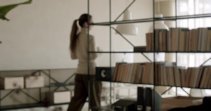 Young woman with long hair in a ponytail walking up the stairs inside a modern home with a large library bookshelf, filmed from behind in a beautiful, cinematic slow motion shot