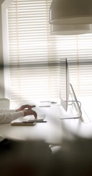 Anonymous person typing on a wireless keyboard at a minimalist white desk in a modern office with natural light coming through the blinds, focusing on productivity and digital workflow. vertical