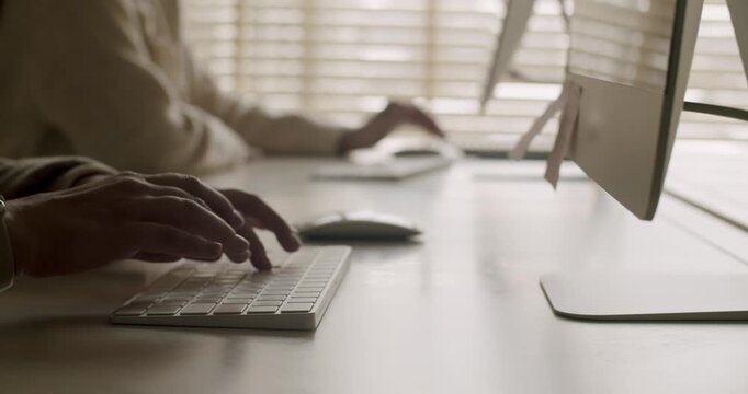 Anonymous office workers typing on wireless keyboards while sitting at a white desk in a contemporary workspace with natural light coming through the blinds