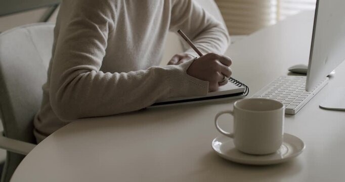Anonymous woman sitting at a modern white desk and taking notes with a pencil in a spiral notebook, studying or working remotely on a computer with a cup of coffee nearby, slow motion