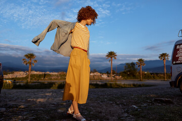 Obraz premium Young woman with curly hair twirls as she throws a light jacket over her striped top in a sunlit desert landscape, conveying movement, spontaneity, and carefree style.