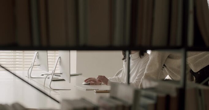 Slow motion view through a bookshelf of a young woman's hands using a computer mouse while sitting at a white desk in a modern office with natural light coming through the blinds