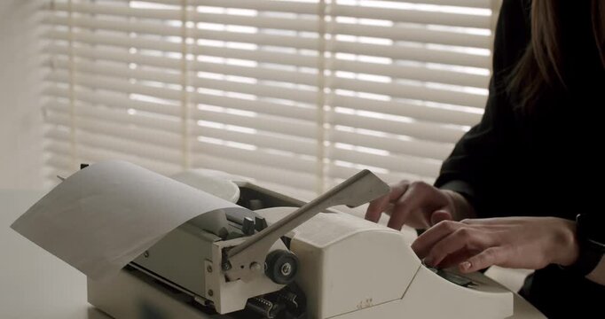 Female journalist's hands typing on a vintage typewriter, creating a new story on a piece of paper in a dimly lit office with blinds in the background, representing old-fashioned journalism