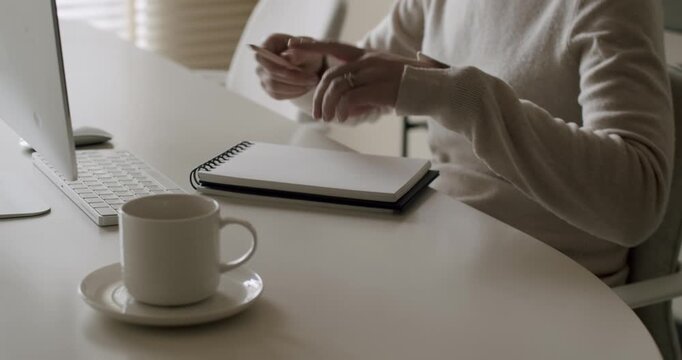Anonymous woman sitting at a modern white desk next to a computer, opening a spiral notebook and beginning to write with a pencil, a cup of coffee resting nearby in a bright minimalist office