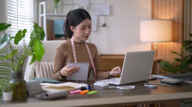 Young Asian fashion designer reviewing sketches while working on laptop at home studio desk