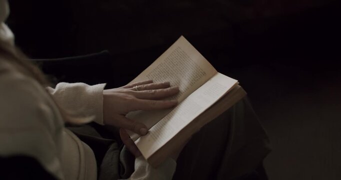 Close-up view of a woman sitting in a dark room and reading an old book with yellowed pages, her fingers gently turning the pages as she becomes engrossed in the story, enjoying a quiet moment