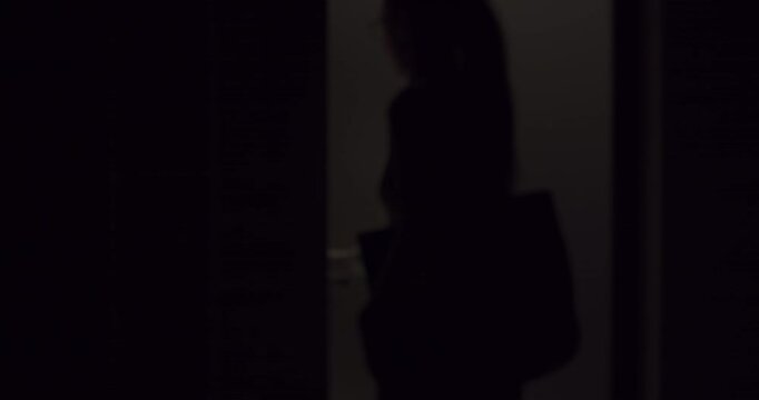 Woman with a ponytail and a tote bag entering a dark apartment at night, her hand touching the wall searching for the light switch before closing the door behind her in slow motion