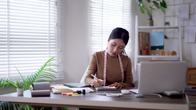 Asian fashion designer sketching garment designs with measuring tape at bright studio workspace