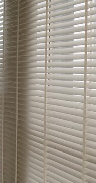 White horizontal blinds on a window moving slowly in a gentle draft, creating a calm and relaxing mood. Subtle sunlight filters through the slats, creating a soft and minimalist background. vertical
